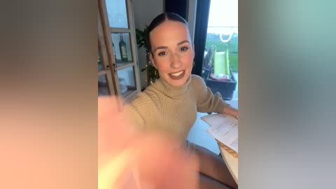 Media: A video of a smiling, light-skinned woman with a ponytail in a beige sweater, seated at a table in a sunlit room with plants and a window.