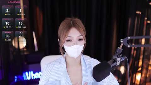 Media: Video of an East Asian woman with light skin and brown hair, wearing a blue-striped blouse, white mask, and glasses, using a microphone on a livestream. Background shows dark curtains and blurred studio elements.