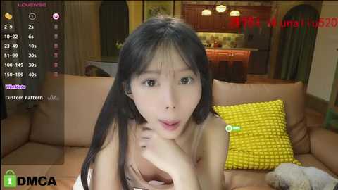 Media: A video of an Asian woman with long black hair, wearing a sleeveless top, in a cozy living room with a yellow cushion and kitchen in the background.