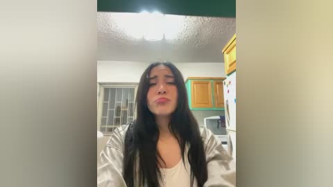 Media: Video of a young woman with long black hair and fair skin, wearing a white shirt and silver jacket, standing in a kitchen with yellow cabinets, green walls, and a white ceiling light.