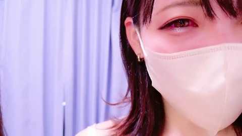 Media: Video of an Asian woman with long black hair, wearing a beige surgical mask, gazing slightly to the right, with soft, even lighting and a blurred blue curtain in the background.