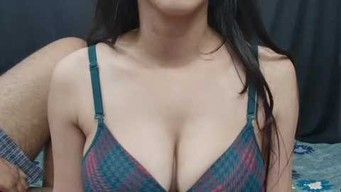 Media: Video of a topless woman with long, straight black hair and medium skin tone, wearing a low-cut, patterned bra. A man's arm with a tattoo is visible in the background.