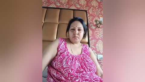 Media: Video of an overweight, middle-aged South Asian woman with dark hair, wearing a sleeveless, patterned pink blouse, seated on a beige bed with a floral wallpaper background.