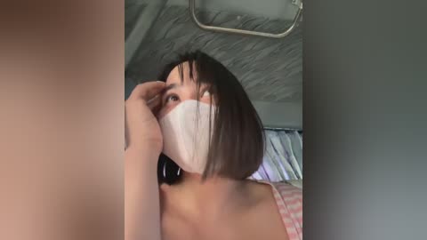 Media: A video of an Asian woman with a white face mask, short bob haircut, and bare shoulders, sitting in a bedroom with a slanted ceiling and a bed with striped sheets.