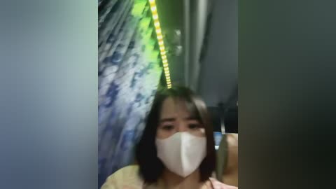 Media: A blurred video of an Asian woman with medium skin tone and straight black hair, wearing a white face mask, standing in a dimly lit corridor with blue and white patterned curtains and green LED lights.