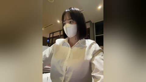 Media: Video of a woman with shoulder-length dark hair, wearing a white face mask and matching white surgical gown, standing in a dimly lit room with modern furnishings.