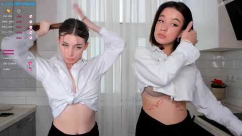 Media: Video of two young women in white shirts, one tying her hair, the other adjusting her earpiece, in a modern kitchen with white tiles and blinds.