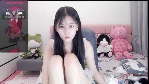 Media: A video of an East Asian woman with long black hair, sitting on a bed with stuffed toys, including a pink bear, in a bedroom.