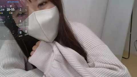 Media: A video shows an Asian woman with long dark hair, wearing a white face mask and a pink sweater, with a digital clock displaying 20:55 on a wall behind her.