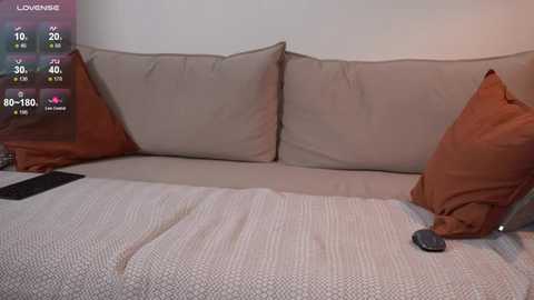 Media: Video of a beige couch with two large, textured pillows and a textured blanket, with a smartphone placed on the right side. The background shows a minimalistic, off-white wall with a digital weather overlay on the left.