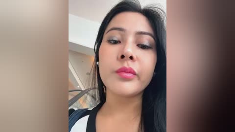 Media: Video of a young woman with long black hair, wearing makeup and a black top, standing in a modern interior with a glass staircase and beige walls.