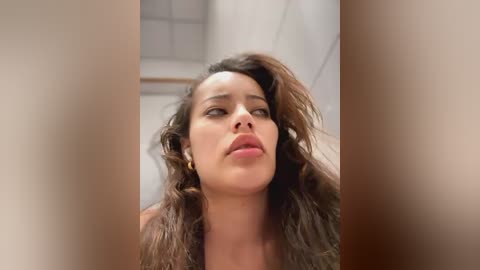 Media: Video of a woman with long, wavy brown hair, wearing gold hoop earrings, and light pink lipstick, looking upward, in a bathroom with a white tiled wall and a grid window in the background.