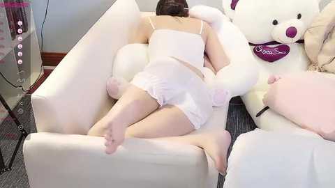 Media: Video of a woman in white lingerie and white gloves lying on a white couch, surrounded by plush toys, including a large teddy bear.