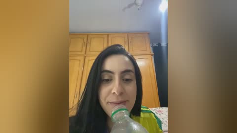 Media: Video of a young woman with long black hair, light skin, and dark eyes, drinking from a green bottle in a bedroom with wooden cabinets and a ceiling fan.