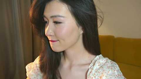 Media: Video of a young Asian woman with long, straight black hair, fair skin, and red lipstick, wearing a floral-patterned blouse, sitting indoors with a beige wall and mustard-colored couch in the background.