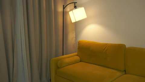 Media: Video of a modern, cozy living room corner featuring a mustard-yellow velvet sofa and a tall, black floor lamp with a white, rectangular shade casting a soft, warm glow. The background includes beige curtains.