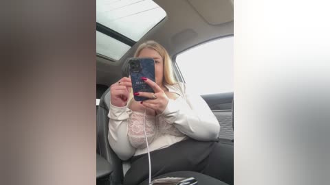 Media: Video of a blonde woman with a large breast in a white lace bra, taking a selfie in a car, holding a smartphone with a pink case, revealing her cleavage.