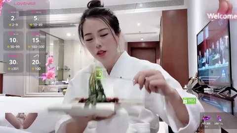 Media: Video of an Asian woman with dark hair in a bun, wearing a white chef's uniform, holding a plate of food in a modern, well-lit kitchen with pink flowers, and a television displaying \"Weedle\" on the right.