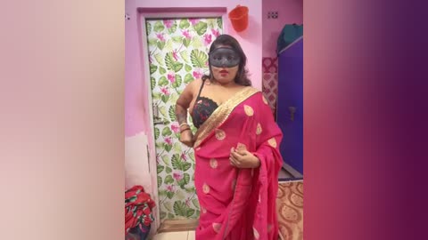 Media: Video of a plus-sized South Asian woman in a pink sari with gold embroidery, black bra, and black mask, standing in a colorful room with floral curtains, blue furniture, and a red suitcase.