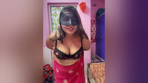 Media: Video of a curvy, light-skinned woman with medium-length brown hair, wearing a black lace bra and a red sari with gold embroidery, standing in a narrow hallway with pink walls, a flowered door, and a laundry basket.