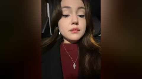 Media: Video of a young woman with long, wavy brown hair, wearing a burgundy top and a black blazer, seated in a dimly lit car. She has a neutral expression, with a silver necklace visible.