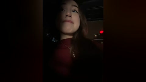 Media: A dimly lit video of a young Asian woman with long brown hair, wearing a burgundy top, looking upwards with a contemplative expression, in an indoor setting with a blurred background.