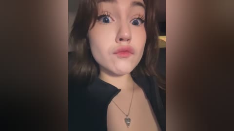 Media: A close-up video of a young woman with fair skin, brown hair, and blue eyes, puckering her lips in a playful kiss. She wears a black jacket and a silver necklace. The background is out of focus.