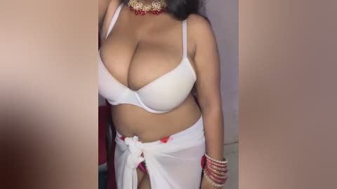 Media: Video of a South Asian woman with medium skin tone, wearing a white bra and a sheer white sarong tied at the waist, adorned with red and gold jewelry, standing indoors.