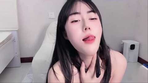 Media: Video of an Asian woman with long black hair, fair skin, and red lipstick, sticking her tongue out, wearing a black strapless top, sitting on a white bed in a minimalist room with a white wall, a white cabinet, and a white toilet.