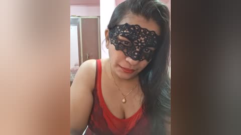 Media: Video of an Asian woman with long dark hair, wearing a black lace masquerade mask covering her eyes and a red tank top. She is indoors, with blurred background elements.