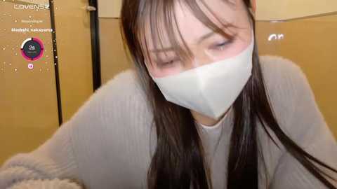 Media: A video of a young Asian woman with long dark hair, wearing a white mask, a grey sweater, and a light blue shirt, blurred in a bathroom with a yellow wall and a digital thermometer showing 21\u00b0C.