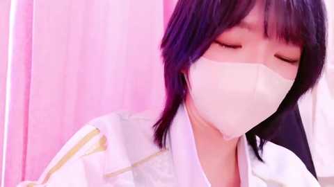 Media: Video of a young Asian woman with shoulder-length black hair, wearing a white surgical mask and lab coat, standing in a pink-lit room.