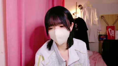 Media: A video of a young Asian woman with straight black hair and a white face mask, wearing a light-colored jacket, standing indoors near a pink curtain and a hanging white coat.