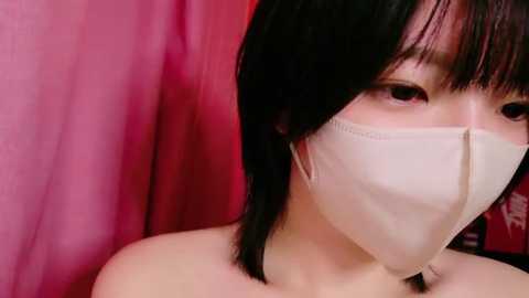 Media: A video of an Asian woman with short black hair and a light beige face mask, against a backdrop of pink and red curtains.