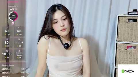 Media: A video of a young East Asian woman with long black hair, wearing a sheer beige top, sitting against a white curtain background.