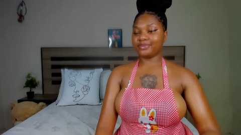 Media: Video of a smiling, dark-skinned Black woman with braided hair, wearing a red-checkered apron, standing in a simple bedroom with a bed and white pillows.