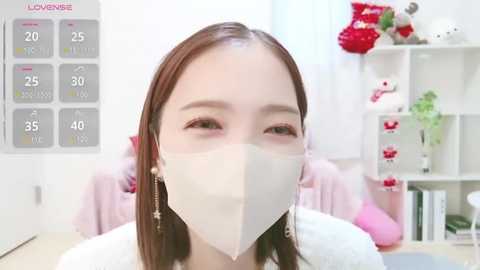 Media: A video of an East Asian woman with straight brown hair, wearing a white face mask, pink shirt, and pearl earrings, in a modern, well-lit room with white shelves, plants, and a calendar.