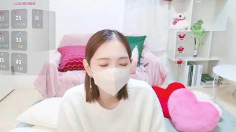 Media: Video of an Asian woman with shoulder-length brown hair wearing a white mask, seated on a light-colored rug in a cozy, pastel-colored room.