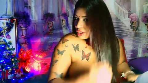 Media: Video of a topless woman with long, straight dark hair, adorned with butterfly tattoos, standing in a grand, decorative room with a floral centerpiece.