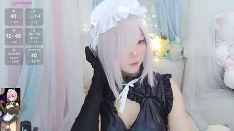 Media: Video of a pale-skinned woman with platinum blonde hair in a white maid headband, wearing a black corset and gloves, sitting in a soft, pastel-colored room.