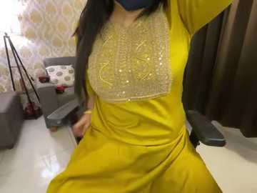 Media: Video of a South Asian woman in a vibrant yellow kurta with intricate embroidery, sitting in a modern living room with a gray couch and patterned wallpaper.