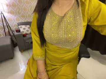 Media: Video of a woman with long black hair wearing a mustard yellow kurta with intricate embroidery and a grey dupatta draped over her shoulders. The background features a modern living room with grey furniture, a patterned wallpaper, and a floor lamp.