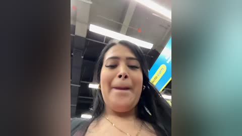 Media: Video of a smiling Latina woman with long black hair, wearing a black top, standing in a brightly lit store with fluorescent lights and blue signage in the background.