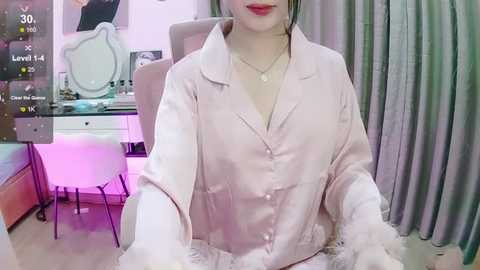 Media: Video of an Asian woman with pale skin, wearing a pink satin robe with white fur trim, sitting in a modern, pink-lit bedroom with a vanity table and grey curtains.