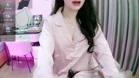 Media: Video of a young woman with long black hair, fair skin, and red lipstick, wearing a light pink button-up shirt, seated in a modern, pink-lit room with a mirror and grey curtains.