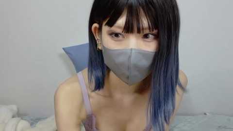 Media: A video of an East Asian woman with long, straight black hair, wearing a light purple spaghetti strap top, gray face mask, and earrings. She has a pale complexion and is indoors, likely in a bedroom, with a white wall and a bed with white sheets in the background.