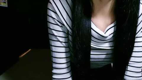 Media: Video of a woman with long black hair, wearing a black-and-white striped shirt, sitting in a dimly lit room with a dark background.