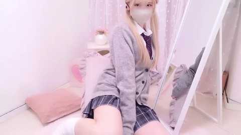 Media: Video of a young Asian woman with long blonde hair in a light grey cardigan, black plaid skirt, white socks, and face mask, kneeling on beige carpet in a softly lit room with a mirror.