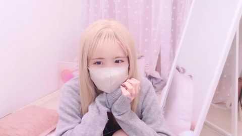 Media: Video of a young girl with long blonde hair, wearing a white face mask and a gray sweater, sitting on a bed with a pink quilt, looking into a mirror. Pastel-colored room with polka-dot curtains.