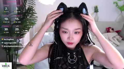 Media: A video of an East Asian woman with fair skin, wearing black cat ears and a sleeveless black top, adjusting her hair in a cozy, brightly-lit room with a plush couch.